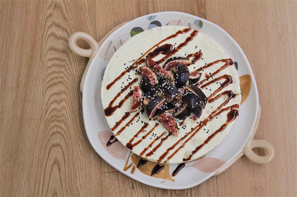 Picture is an overhead shot of a round cream-coloured cheesecake, piled cut figs and drizzle with dark date syrup.