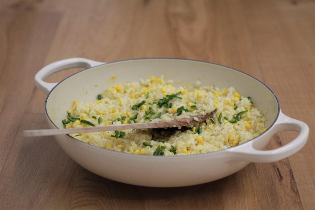 Image shows a cream coloured shallow pot filled with corn risotto garnished with chopped dark green chilli, with a wooden spoon.