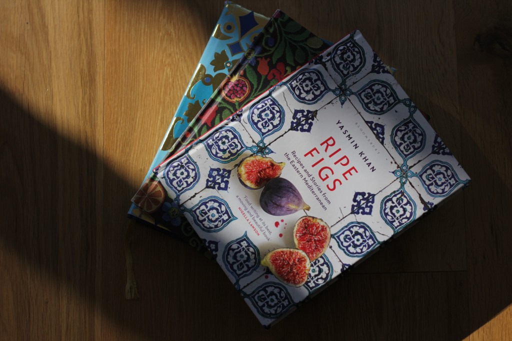 A shaft of light falls on a stack of vibrant decorated cookbook covers. The top book is white with an elaborate blue tile pattern, photos of cut figs, and the title 'Ripe Figs' Yasmin Khan.