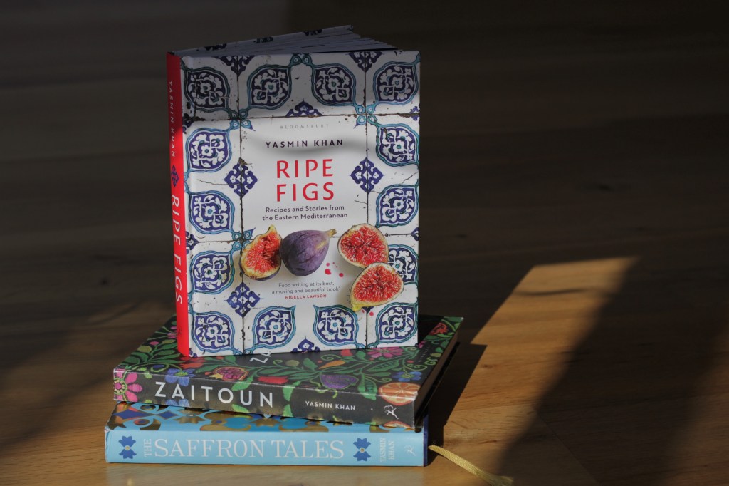 A shaft of light falls on a stack of vibrant decorated cookbooks. The top book is white with an elaborate blue tile pattern, photos of cut figs, and the title 'Ripe Figs' Yasmin Khan.