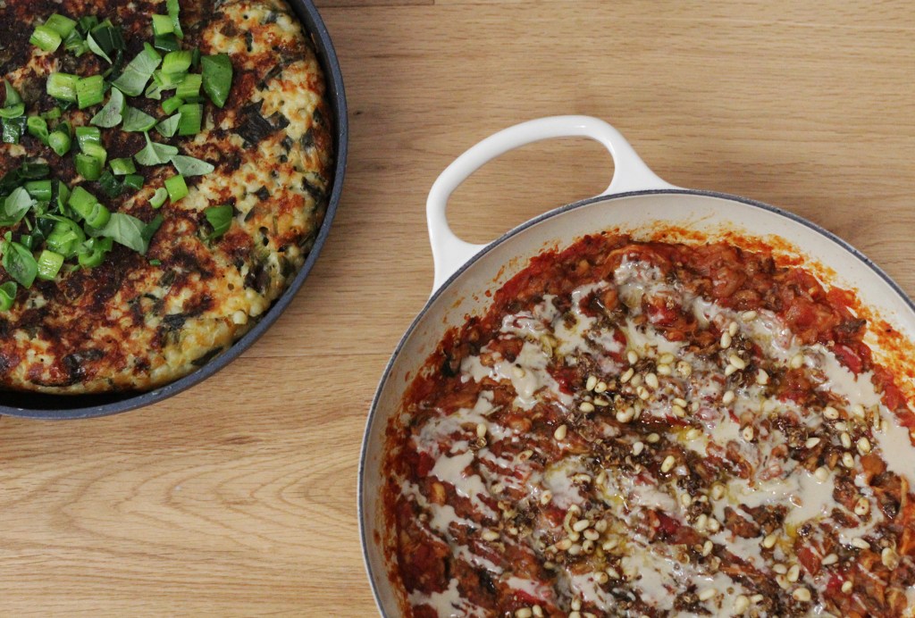 Overhead view of two frying pans, one with a golden brown frittata-style cake topped with vibrant green spring onions and chopped basil, the other with a rich red and brown sauce drizzled in glossy, creamy tahini and dotted with pine nuts.