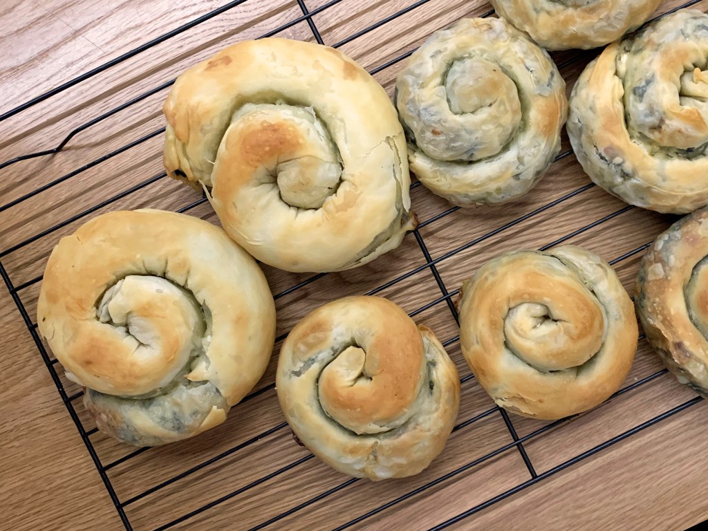 Image shows a baking tray laden with individual coils of spanakopita, in varying sizes and varying degrees of flaky, golden exterior.