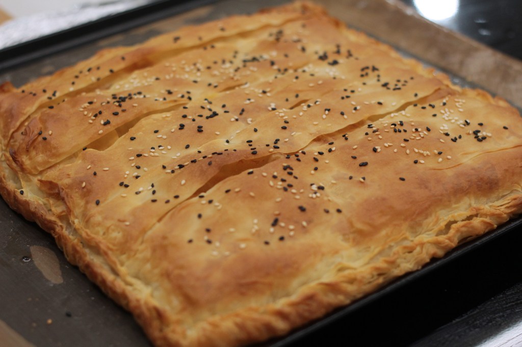 Image shows a square pie on a flat baking sheet, baked to golden perfect with hand-crimped edges and black and white sesame seeds scattered on top.