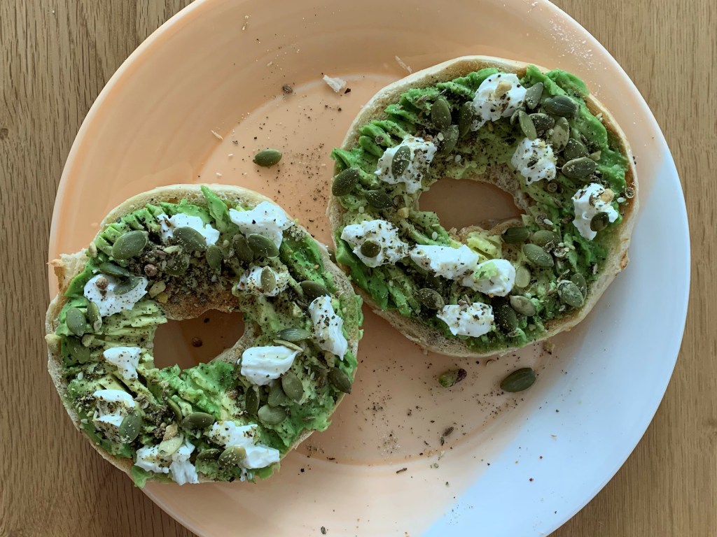 Image shows an open bagel with smashed avocado on both halves, scattered with dukkah, dollops of goat cheese, and pepitas.