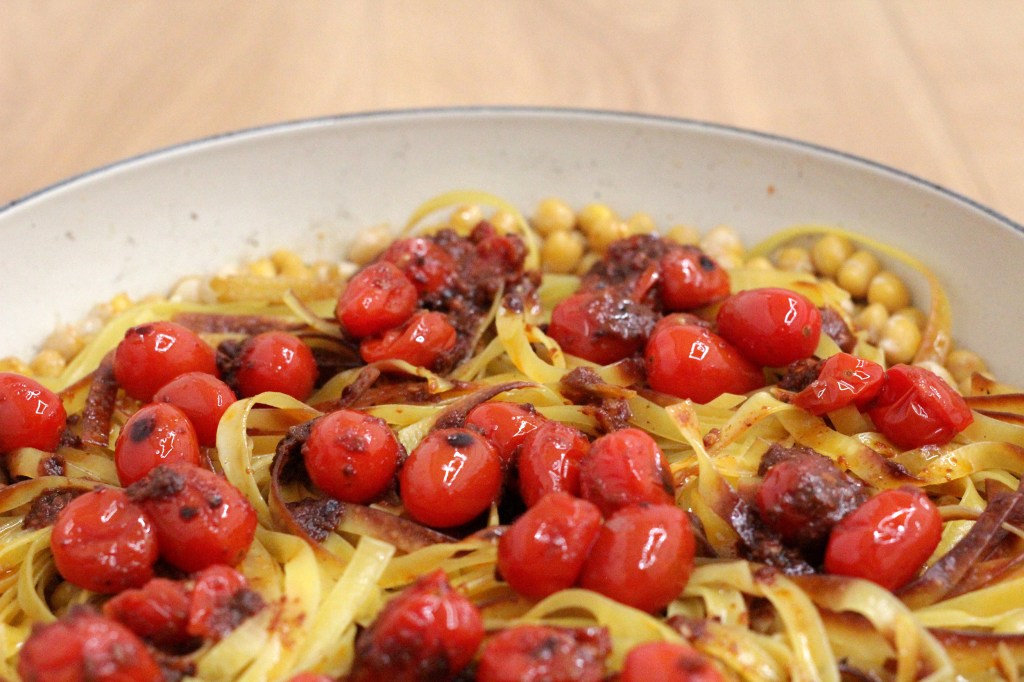 A close up of a cooking dish fill with golden yellow tagliatelle, chickpeas and charred red cherry tomatoes.